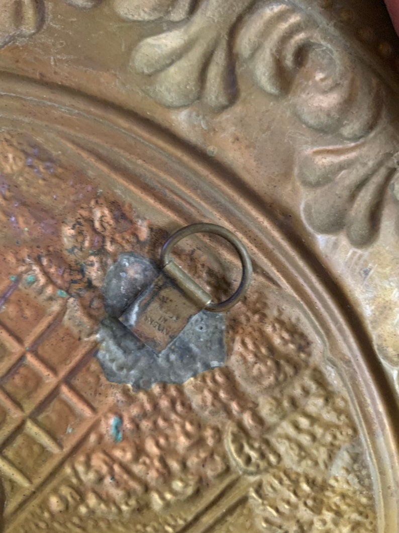 K&ouml;nnte beinhalten: Nahaufnahme einer goldfarbenen Wanddekoration aus Metall mit einem dekorativen Design. Die Dekoration hat eine Metallschaufel auf der R&uuml;ckseite zum Aufh&auml;ngen.