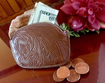May include: A brown leather coin purse with a floral embossed design. The purse is open and contains a twenty dollar bill and several copper pennies.