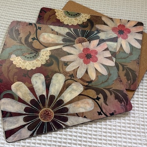 May include: A set of three rectangular placemats featuring a floral pattern with large white petals, dark brown centers, and swirling brown and green foliage accents. Each placemat has a smooth top surface and a brown cork backing for stability.