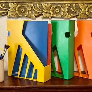 May include: Three colorful plastic magazine holders, one yellow, one green, and one orange, are arranged on a wooden surface. The holders are designed with a unique geometric pattern and are ideal for organizing and storing magazines, papers, or other documents.