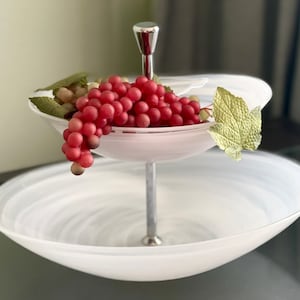 Può includere: Un portavivande bianco a due livelli con una base in metallo argentato e un singolo grappolo di uva rossa sul livello superiore.