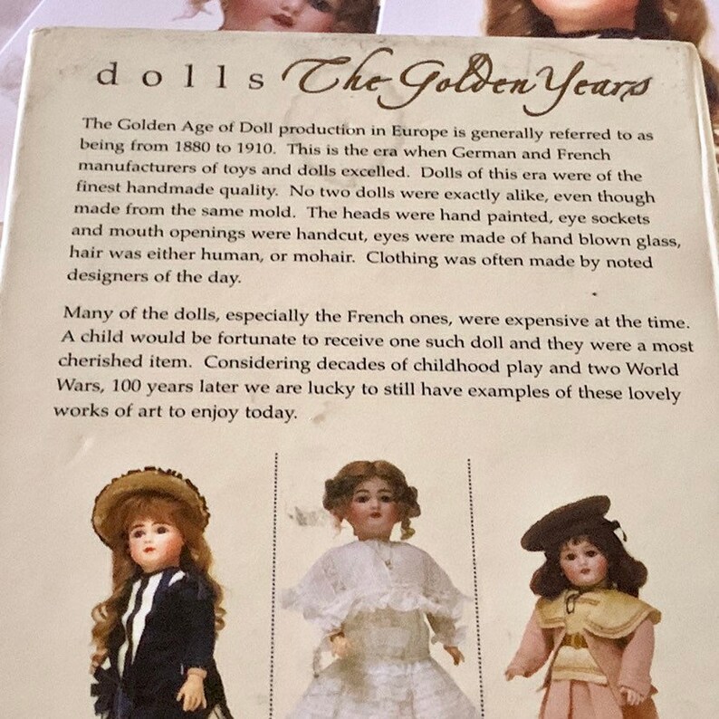 Puede incluir: Una portada de libro con el t&iacute;tulo "Dolls The Golden Years" y un extracto de texto sobre la historia de la fabricaci&oacute;n de mu&ntilde;ecas en Europa desde 1880 hasta 1910. La portada del libro presenta tres mu&ntilde;ecas antiguas con rostros de porcelana y ropa elaborada.