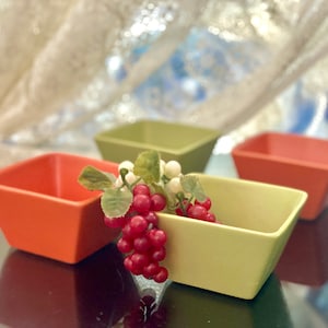 Può includere: Tre ciotole quadrate in ceramica di colore arancione, verde e giallo. Le ciotole sono disposte in fila con un rametto di bacche rosse e fiori bianchi nella ciotola centrale.