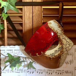 May include: A red, white and gold masquerade mask with a cracked, distressed finish. The mask has a single eye hole and is decorated with black swirls and musical notes.