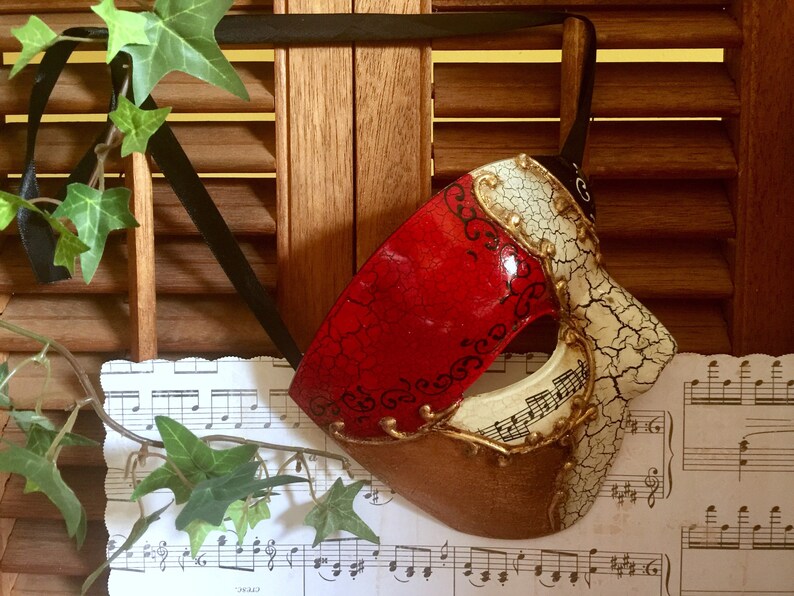 May include: A red, white and gold masquerade mask with a cracked, distressed finish. The mask has a single eye hole and is decorated with black swirls and musical notes.