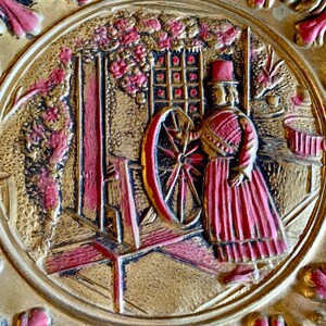 K&ouml;nnte beinhalten: Eine goldene und rote Reliefplatte, die eine Frau in einem roten Kleid und Hut zeigt, die ein Spinnrad bedient. Die Platte ist vor einem Hintergrund aus stilisierten Blumen und Ranken.