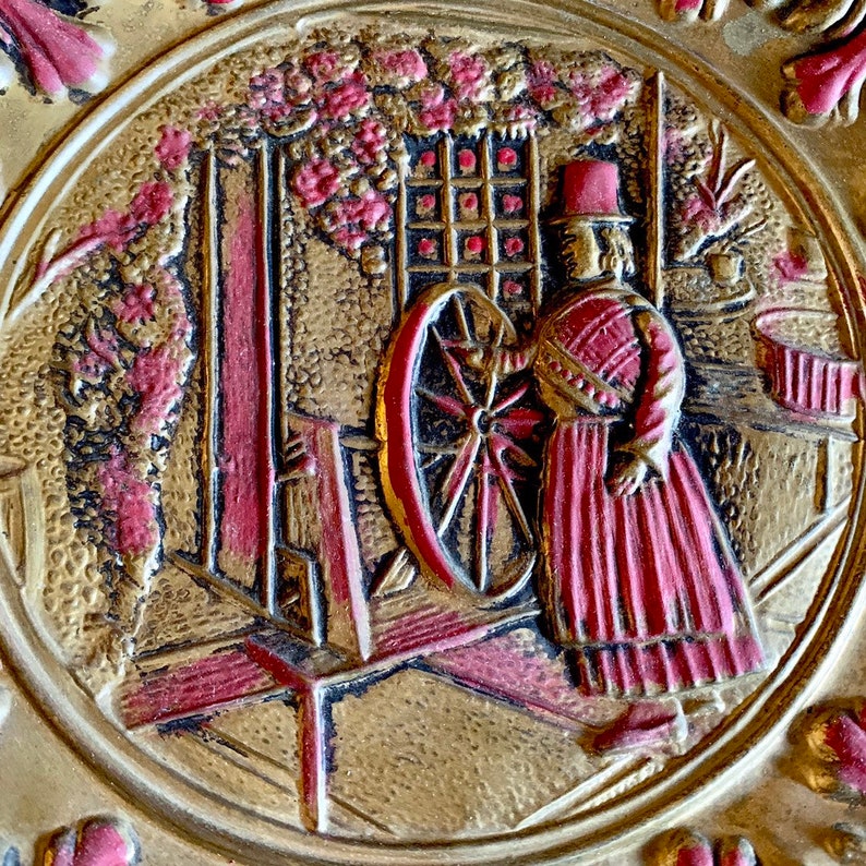 K&ouml;nnte beinhalten: Eine goldene und rote Reliefplatte, die eine Frau in einem roten Kleid und Hut zeigt, die ein Spinnrad bedient. Die Platte ist vor einem Hintergrund aus stilisierten Blumen und Ranken.