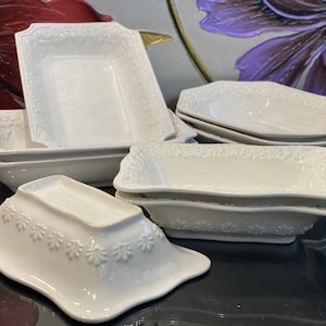 May include: A set of white ceramic serving dishes with a floral design. The dishes are various sizes and shapes, including rectangular and oval.