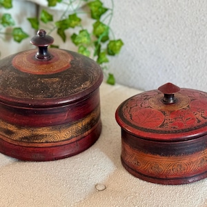 Pode incluir: Duas caixas redondas de madeira com tampas. A caixa maior é vermelha com desenhos dourados e pretos. A caixa menor é vermelha com desenhos florais dourados e pretos.