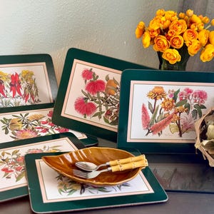 Peut inclure: Ensemble de sets de table floraux avec une bordure verte et un intérieur crème. Chaque set de table présente une illustration botanique différente. Un plat en forme de feuille avec des couverts à manche en bambou est posé sur un set de table. Un vase de roses jaunes est en arrière-plan.