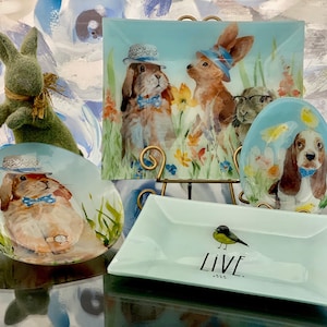 Puede incluir: Cuatro platos de vidrio decorativos con un tema primaveral. Los platos presentan conejos, perros y pájaros con sombreros y pajaritas. Los platos están sobre una superficie negra con un soporte de metal dorado.