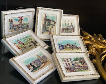 May include: Set of seven small framed watercolour paintings depicting various cityscapes. Each painting is framed in a white frame with a gold trim. The paintings are arranged in a stack on a black surface.