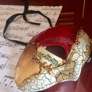 May include: A red, gold and white masquerade mask with a cracked white finish and black accents. The mask has a musical note design and a black ribbon.