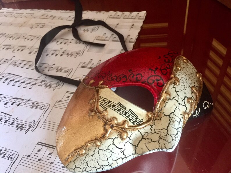 May include: A red, gold and white masquerade mask with a cracked white finish and black accents. The mask has a musical note design and a black ribbon.