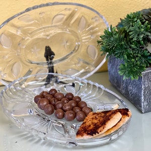 Può includere: Un piatto da portata in vetro trasparente con un motivo decorativo. Il piatto è pieno di noci di macadamia ricoperte di cioccolato e due pezzi di pane tostato.