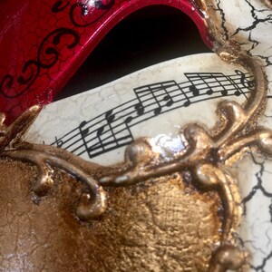 May include: A red and gold masquerade mask with a white background and musical notes printed on it. The mask has a decorative gold design around the edges.