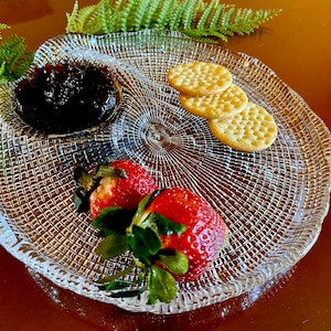 Pode incluir: Um prato de servir de vidro transparente e texturizado com três compartimentos. Uma seção contém geleia escura, outra tem três biscoitos e a última contém dois morangos vermelhos. Samambaias ao fundo.