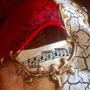 May include: A masquerade mask with a red, white and gold design. One side of the mask features a red background with black swirls and the other side features a white background with gold musical notes and a cracked effect.
