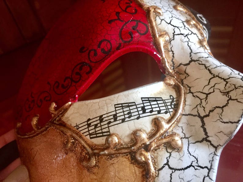 May include: A masquerade mask with a red, white and gold design. One side of the mask features a red background with black swirls and the other side features a white background with gold musical notes and a cracked effect.