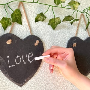 May include: Two black heart-shaped chalkboard signs hanging on a white wall with green ivy vines. The sign on the left has the word "Love" written on it in white chalk. A hand is holding a piece of chalk and writing on the sign.