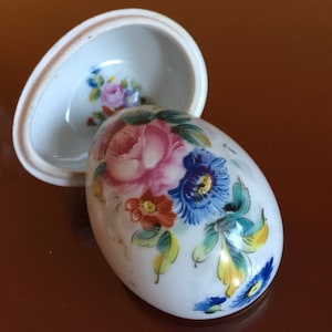 May include: A white porcelain egg-shaped trinket box with a floral design. The lid is open and shows the inside of the box. The flowers are painted in shades of pink, blue, yellow, and orange.