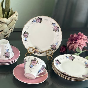 May include: A set of floral patterned teacups and saucers. The cups and saucers are white with pink, blue, and black floral designs. The saucers are a pale pink color. The set is displayed on a dark surface.