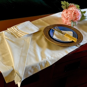 May include: A white table runner with a set of folded napkins and a place setting with a blue plate, a woven placemat, a knife, and a fork. A bouquet of pink flowers is on the table runner.
