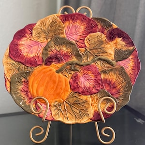 Puede incluir: Un plato de servir de cerámica decorativo con una calabaza y hojas de otoño en tonos marrones, verdes, rojos y dorados. El plato está sobre un soporte de metal dorado.