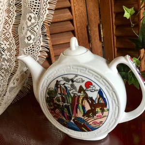 Könnte beinhalten: Weiße Porzellan-Teekanne mit einem farbenfrohen asiatisch inspirierten Design mit Bergen, Bäumen, Vögeln und Hirschen. Die Teekanne hat einen runden Körper und einen geschwungenen Ausguss.