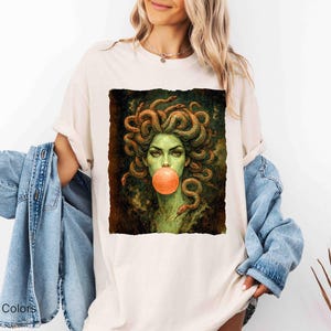 K&ouml;nnte beinhalten: Elfenbeinfarbenes T-Shirt mit einer Grafik von Medusa, dargestellt mit Schlangen als Haare und einer Kaugummiblase. Das Kunstwerk verwendet Gr&uuml;n- und Braunt&ouml;ne. Der Text "Comfort Colors" und "Ivory" ist unten links aufgedruckt.