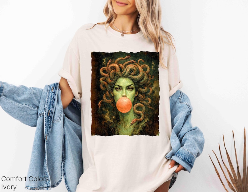 K&ouml;nnte beinhalten: Elfenbeinfarbenes T-Shirt mit einer Grafik von Medusa, dargestellt mit Schlangen als Haare und einer Kaugummiblase. Das Kunstwerk verwendet Gr&uuml;n- und Braunt&ouml;ne. Der Text "Comfort Colors" und "Ivory" ist unten links aufgedruckt.
