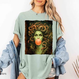 K&ouml;nnte beinhalten: Hellgr&uuml;nes T-Shirt mit einer Medusa-Grafik, die eine Kaugummiblase bl&auml;st. Das Kunstwerk zeigt Medusa mit gr&uuml;ner Haut, Schlangenhaaren und einer kaugummifarbenen Blase. Der Text "Comfort Colors Bay" ist unten links aufgedruckt.