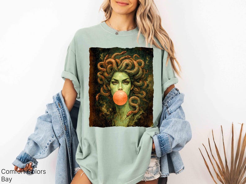 K&ouml;nnte beinhalten: Hellgr&uuml;nes T-Shirt mit einer Medusa-Grafik, die eine Kaugummiblase bl&auml;st. Das Kunstwerk zeigt Medusa mit gr&uuml;ner Haut, Schlangenhaaren und einer kaugummifarbenen Blase. Der Text "Comfort Colors Bay" ist unten links aufgedruckt.