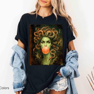 K&ouml;nnte beinhalten: Schwarzes T-Shirt mit einer Medusa-Grafik, die Schlangen als Haare und eine Kaugummiblase zeigt. Das Bild hat eine gr&uuml;ne und orange Farbpalette. Der Text "Comfort Colors Black" befindet sich unten links.