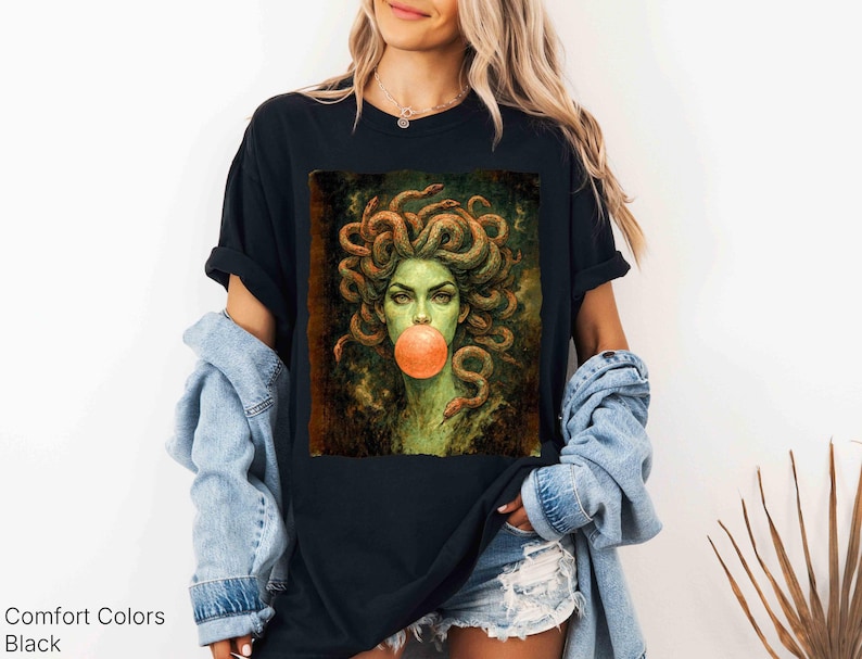 K&ouml;nnte beinhalten: Schwarzes T-Shirt mit einer Medusa-Grafik, die Schlangen als Haare und eine Kaugummiblase zeigt. Das Bild hat eine gr&uuml;ne und orange Farbpalette. Der Text "Comfort Colors Black" befindet sich unten links.