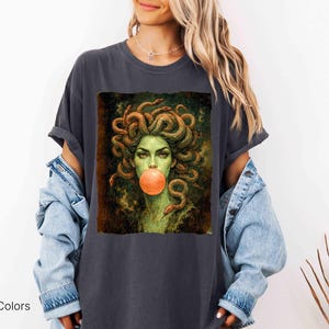 K&ouml;nnte beinhalten: Dunkelgraues T-Shirt mit einem Medusa-Motiv, dargestellt mit Schlangenhaaren und einer Kaugummiblase. Das Design hat eine Vintage-&Auml;sthetik. Der Text "Comfort Colors Pepper" ist unten links aufgedruckt.