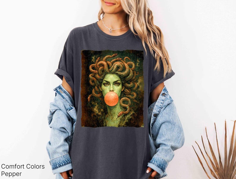 K&ouml;nnte beinhalten: Dunkelgraues T-Shirt mit einem Medusa-Motiv, dargestellt mit Schlangenhaaren und einer Kaugummiblase. Das Design hat eine Vintage-&Auml;sthetik. Der Text "Comfort Colors Pepper" ist unten links aufgedruckt.