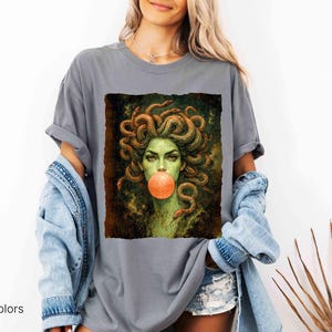 K&ouml;nnte beinhalten: Graues Comfort Colors T-Shirt mit einem Medusa-Motiv. Das Kunstwerk zeigt Medusa mit Schlangen als Haare, einem gr&uuml;nen Teint und einer Kaugummiblase. Das Shirt hat eine entspannte Passform.
