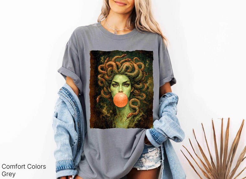 K&ouml;nnte beinhalten: Graues Comfort Colors T-Shirt mit einem Medusa-Motiv. Das Kunstwerk zeigt Medusa mit Schlangen als Haare, einem gr&uuml;nen Teint und einer Kaugummiblase. Das Shirt hat eine entspannte Passform.