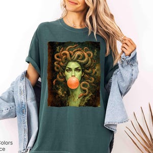 K&ouml;nnte beinhalten: Blaugr&uuml;nes T-Shirt mit einem Medusa-Motiv, das Schlangen als Haare und eine Kaugummiblase zeigt. Die Grafik befindet sich in einem rechteckigen Rahmen. Die Worte "Comfort Colors" und "Blue Spruce" sind unten links aufgedruckt.