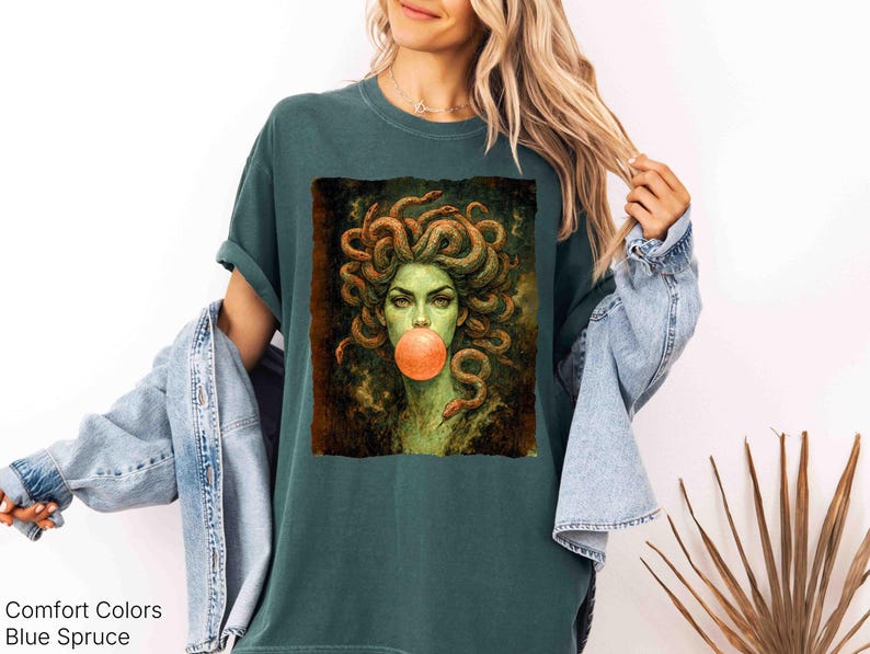 K&ouml;nnte beinhalten: Blaugr&uuml;nes T-Shirt mit einem Medusa-Motiv, das Schlangen als Haare und eine Kaugummiblase zeigt. Die Grafik befindet sich in einem rechteckigen Rahmen. Die Worte "Comfort Colors" und "Blue Spruce" sind unten links aufgedruckt.