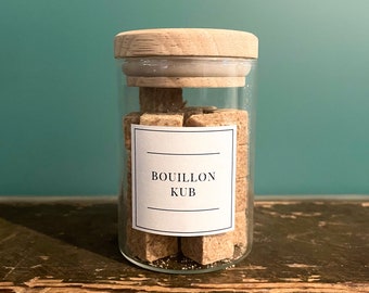May include: A glass jar filled with brown bouillon cubes. The jar has a wooden lid and a white label that reads "BOUILLON KUB".
