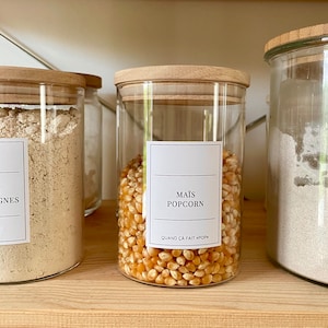 Könnte beinhalten: Ein Glasgefäß mit einem Holzdeckel, gefüllt mit Popcorn-Kernen. Das Gefäß hat ein weißes Etikett mit dem Text "MAÏS POPCORN" und "QUAND ÇA FAIT "POP"".