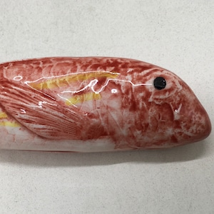 Op de afbeelding: Een keramiek visfiguur met een rode en gele glazuur. De vis heeft een zwart oog en een gedetailleerde textuur.