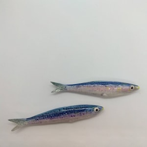 Op de afbeelding: Twee keramische visfiguren, een blauwe en een paarse, met witte en gele accenten. De vissen liggen op een wit oppervlak.