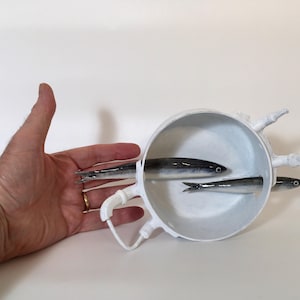 May include: A white ceramic bowl with a handle and three fish sculptures inside. The fish are silver and black, with detailed fins and eyes. The bowl is held by a hand.