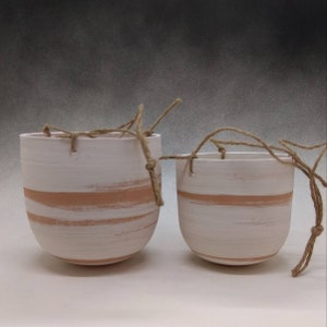 May include: Two white ceramic hanging planters with a light brown stripe design and natural twine hanging cords.