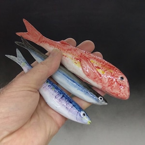 Può includere: Tre figurine di pesce in ceramica. Un pesce rosso con segni gialli e bianchi, e due pesci blu e bianchi più piccoli.