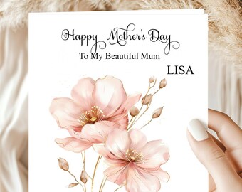 May include: A Mother's Day card with the text "Happy Mother's Day To My Beautiful Mum LISA". The card features watercolor-style pink flowers with gold stems and leaves. The card is held by a person.