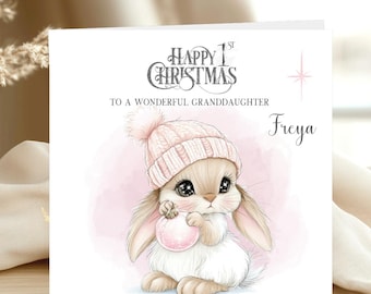 May include: A square greeting card featuring a watercolor illustration of a fluffy bunny wearing a pink knit hat with a pom-pom, holding a pink ornament. The text reads Happy 1st Christmas to a wonderful granddaughter Freya with a pink star.